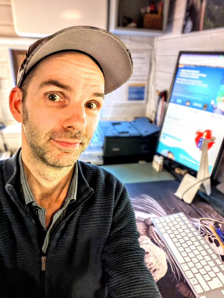 Selfie-Aufnahme von Robert Welti im dunkelblauen Rippstrick-Pullover und mit grauer Kappe an seinem Schreibtisch mit weißer Tastatur, Drucker und einem hochkant ausgerichteten Computermonitor, auf dem eine Webseite zum Praxis-Business geöffnet ist.