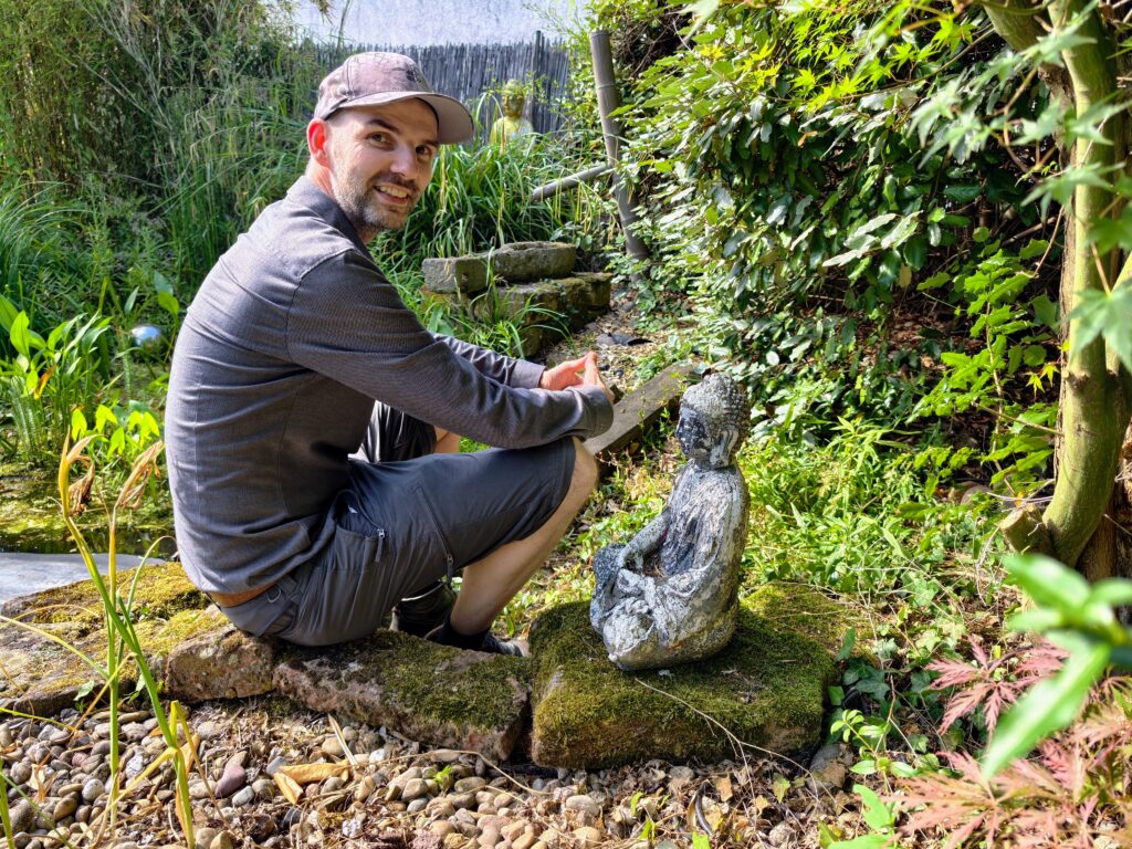 Robert Welti hockt seitlich in grauer Outdoor-Kleidung und mit Kappe auf bemoosten Steinen in einem grünen Garten vor einer kleinen Buddha-Statue aus Stein und lächelt in die Kamera.